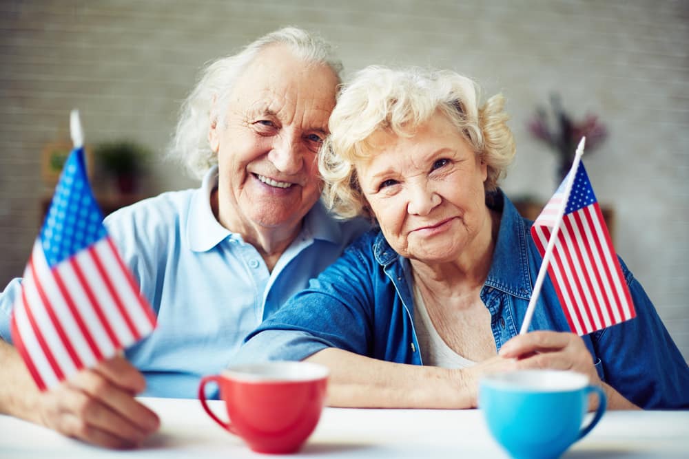 Senior couple holding american flags at kitchen table, mugs in foreground