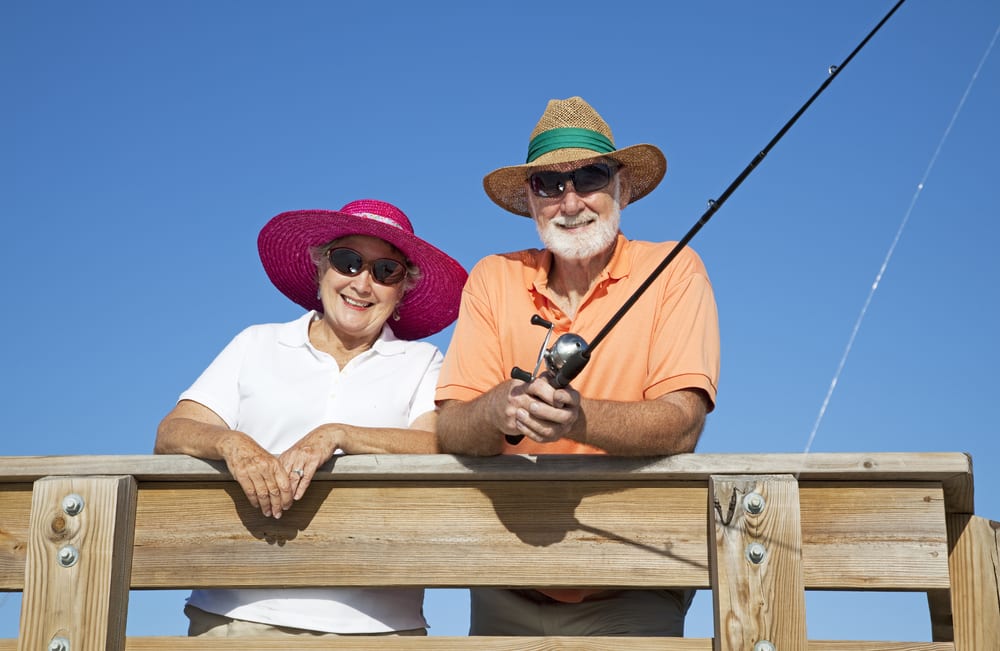 Senior couple wearing sunhats and sunglasses, leading over railing, man fishing